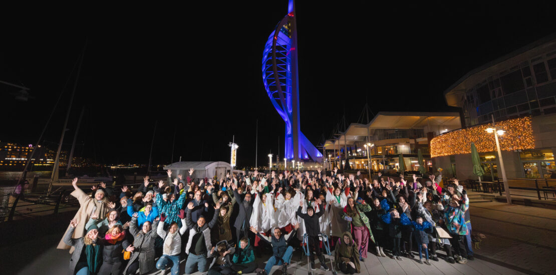 A large crowd of people, cheering with arms up in the air in front of a blue lit Spinnaker Tower.